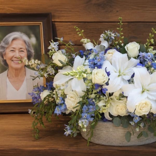 White lilies and roses in a low basket beside a memorial urn and framed portrait