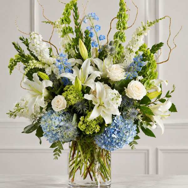 White lilies, roses, blue flowers, and green blooms in a clear glass vase