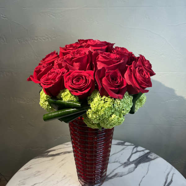Red roses arranged in a tall red glass vase with green accents