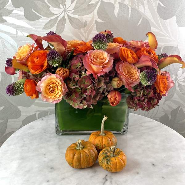 Orange and pink floral arrangement in a square glass vase with small pumpkins