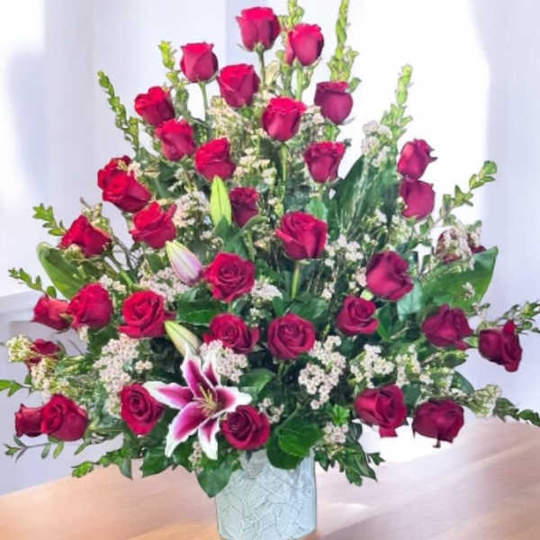 Tall arrangement of red roses with pink lilies and white filler flowers in a white ceramic vase