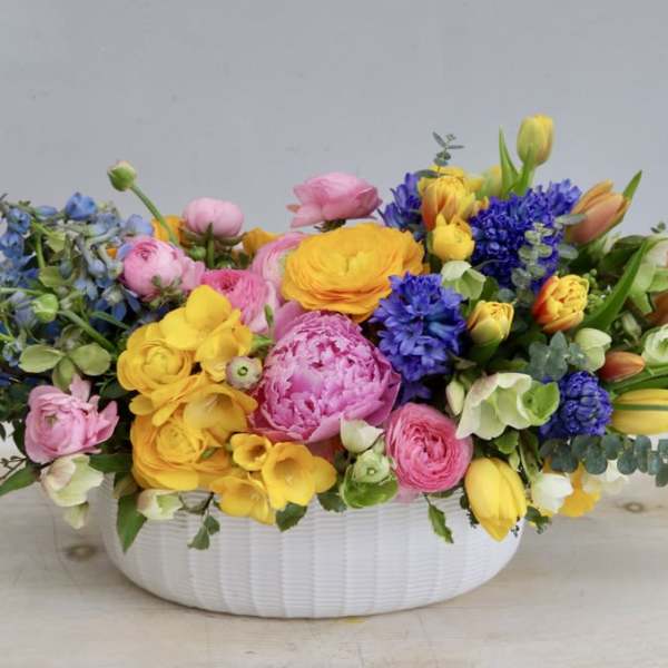 Low white bowl of bright pink, yellow, orange, and blue spring flowers in a lush arrangement