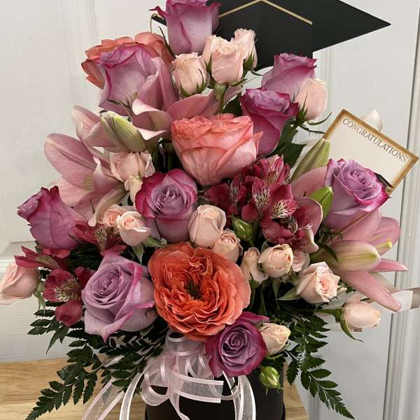 Pink and lavender roses with lilies in a black hatbox, topped with a graduation cap.
