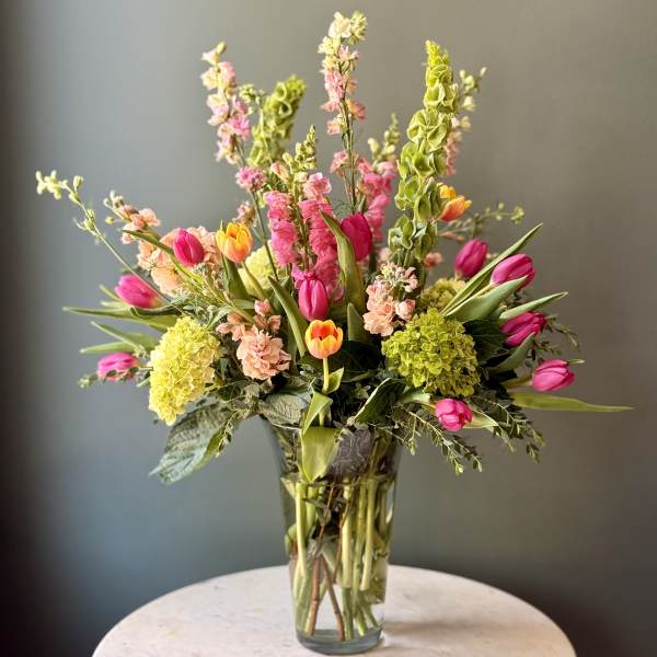 Tall mixed arrangement of pink and yellow tulips, green hydrangeas, and Bells of Ireland in a clear glass vase