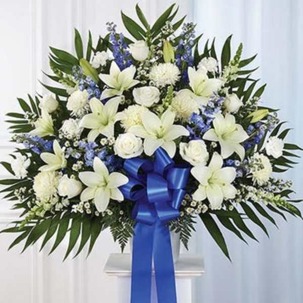 White lilies and roses with blue flowers in a standing spray with a blue ribbon