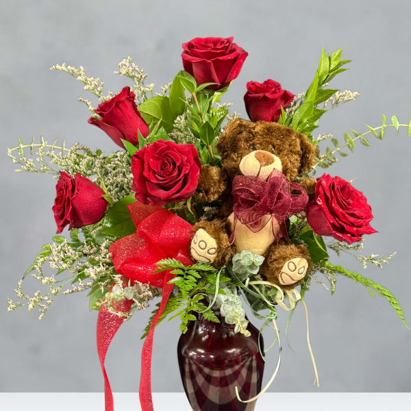 Red roses arranged in a vase with a teddy bear and ribbon accents