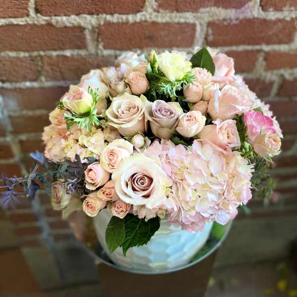 Round arrangement of blush roses and hydrangeas in a white vase against a brick wall.