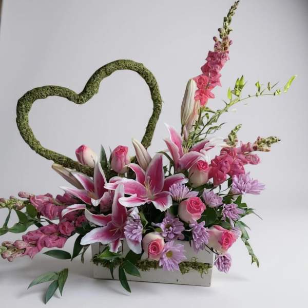 Pink and lavender floral arrangement with a moss heart frame in a white container