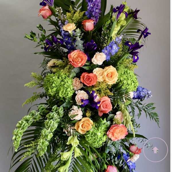 Tall standing arrangement of orange roses, purple delphinium, irises, and green hydrangeas with bold tropical foliage.