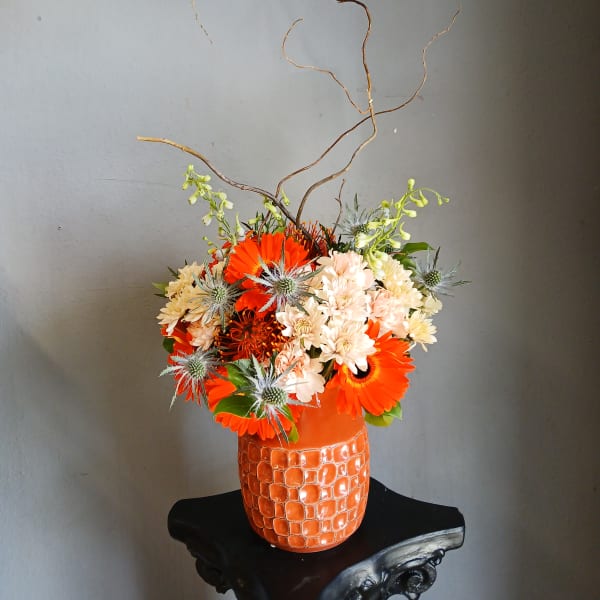 Orange and cream mixed flower arrangement in a textured orange vase with curly branches