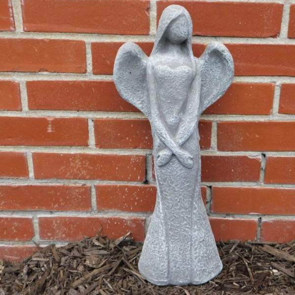 Tall gray stone angel statue with folded arms standing against a red brick wall.