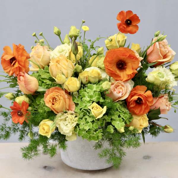Low centerpiece of orange ranunculus, peach and yellow roses, and green hydrangeas in a white pot.