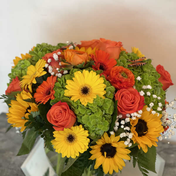Bouquet of yellow daisies, orange roses, and green hydrangeas in a glass vase