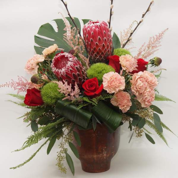 Tropical bouquet with red protea, roses, and carnations in a brown vase