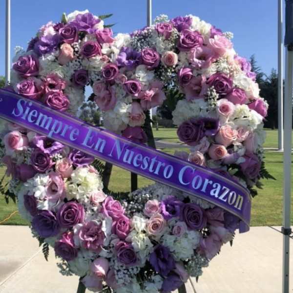 Heart-shaped floral wreath with purple and pink roses and a ribbon reading "Siempre En Nuestro Corazon"