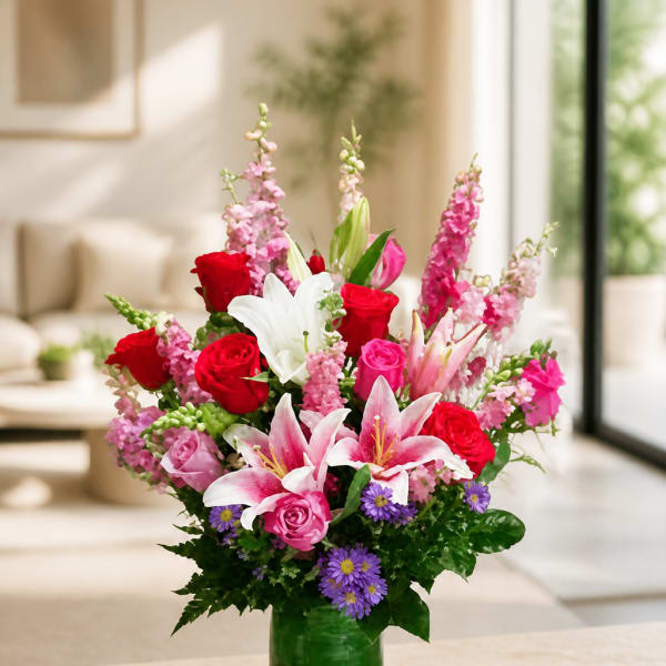 Mixed bouquet of roses, lilies, and purple daisies in a green glass vase
