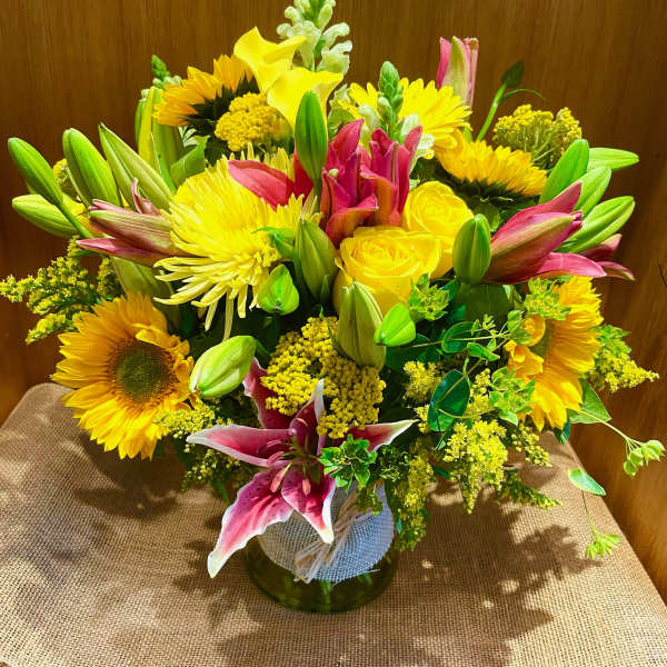 Mixed bouquet with yellow sunflowers, roses, lilies, and pink blooms in a glass vase