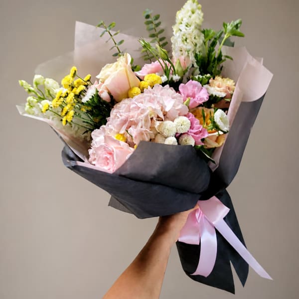 Handheld bouquet of pink, white, and yellow flowers wrapped in dark paper with a pink ribbon