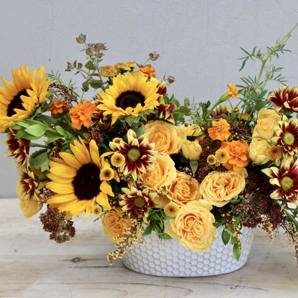 Low oval arrangement of yellow sunflowers, roses, and orange blooms in a white textured ceramic bowl