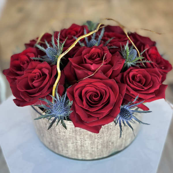 Red roses arranged in a round silver box with blue thistle accents