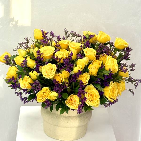 Rounded arrangement of yellow roses with purple filler flowers in a cream ceramic pot