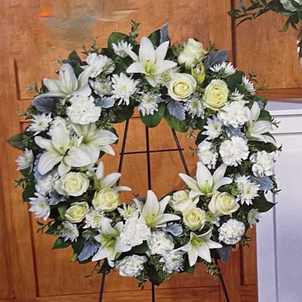 White floral wreath on a black stand with lilies and roses
