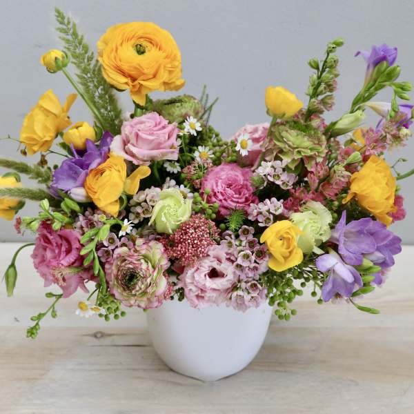 Low arrangement of yellow ranunculus, pink blooms, and purple flowers in a white bowl vase