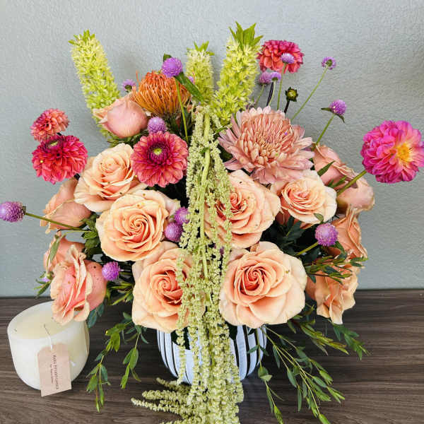 Peach roses and pink blooms arranged in a striped vase with a candle beside it