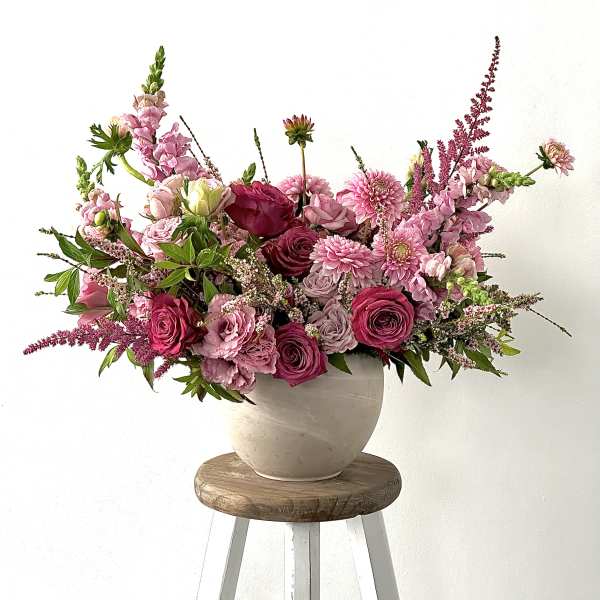Pink mixed flower arrangement in a beige vase on a wooden stool