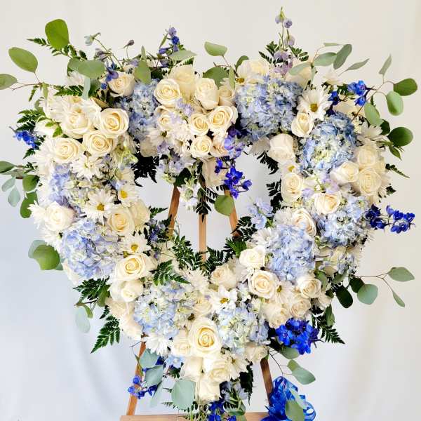 Heart-shaped floral wreath with white roses, blue hydrangeas, and daisies on an easel