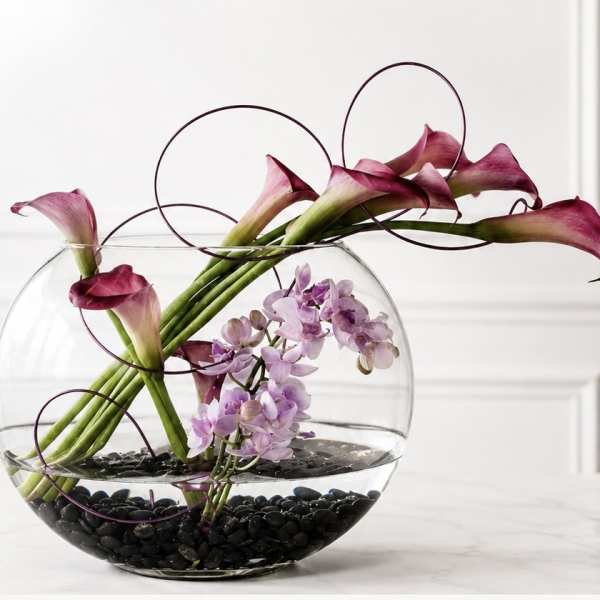 Calla lilies and purple orchids in a round glass bowl with black stones