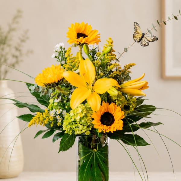 Yellow lilies and sunflowers in a clear glass vase with a butterfly pick
