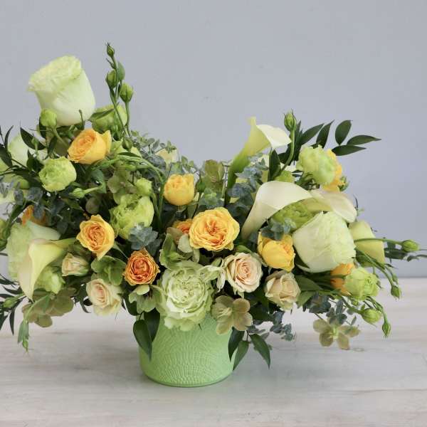 Low yellow and cream flower arrangement with roses and calla lilies in a green ceramic pot