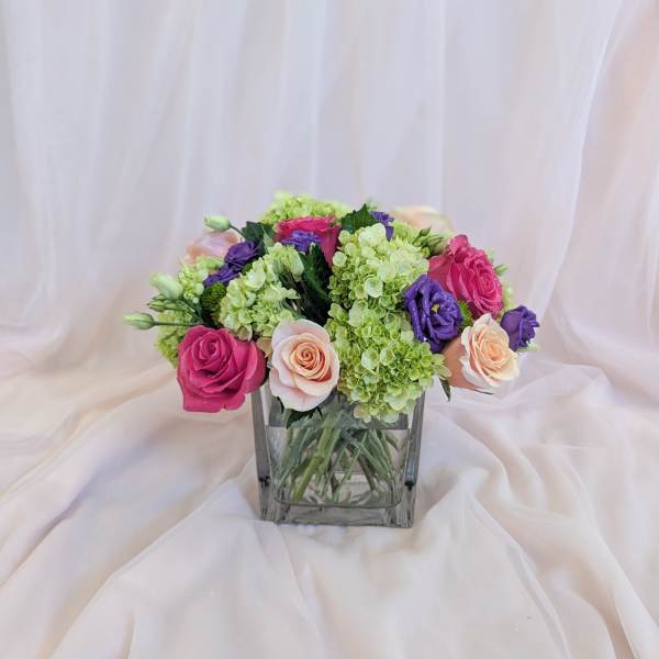 Mixed pink, purple, and green flower arrangement in a square glass vase