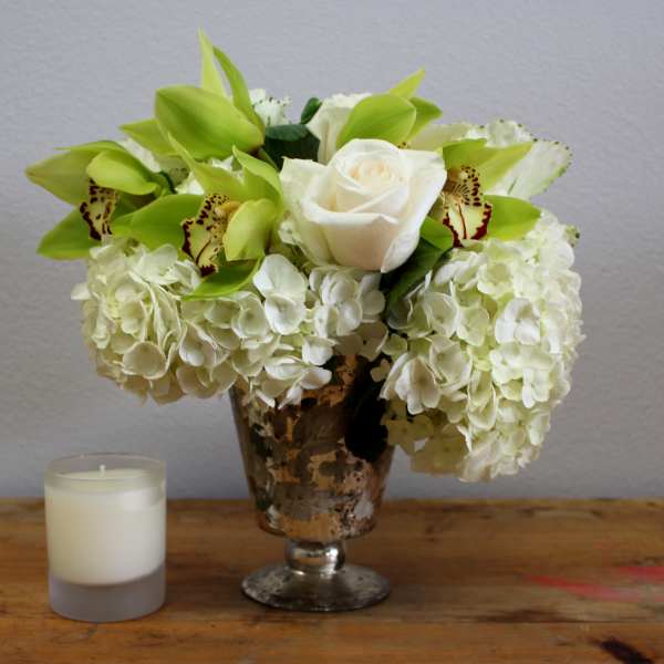 White roses and green orchids in a silver vase beside a candle