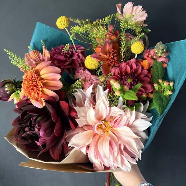 Handheld bouquet of pink, peach, and burgundy flowers wrapped in teal paper
