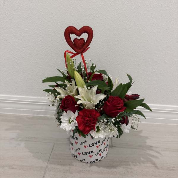 Red and white floral arrangement in a heart-decorated box with a heart topper