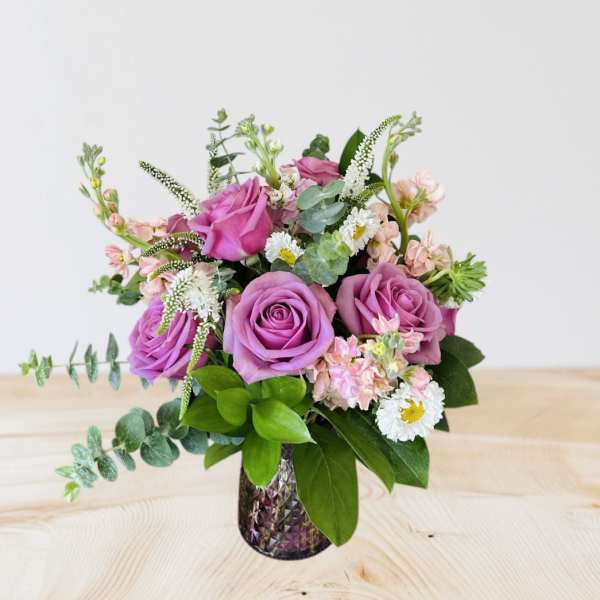 Pink and lavender roses arranged in a textured glass vase with small white and pink accent flowers.