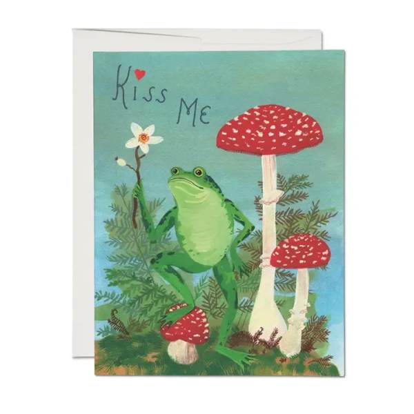 Folded greeting card with a frog holding a white flower among red mushrooms and the words “Kiss Me.”