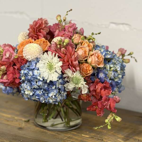 Mixed bouquet of blue hydrangeas, peach roses, and pink flowers in a glass vase