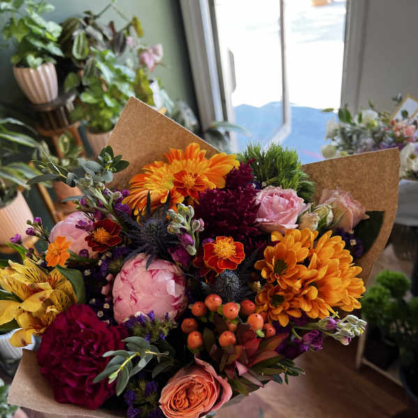 Handheld bouquet of mixed orange, pink, and burgundy flowers in brown paper