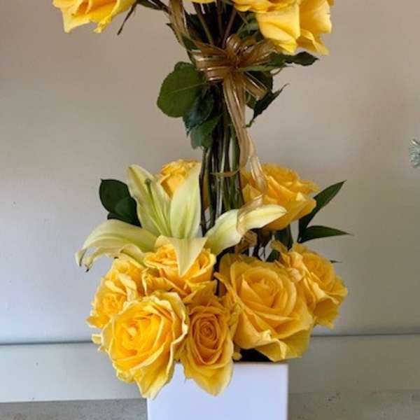 Tall arrangement of yellow roses and pale lilies in a white square vase
