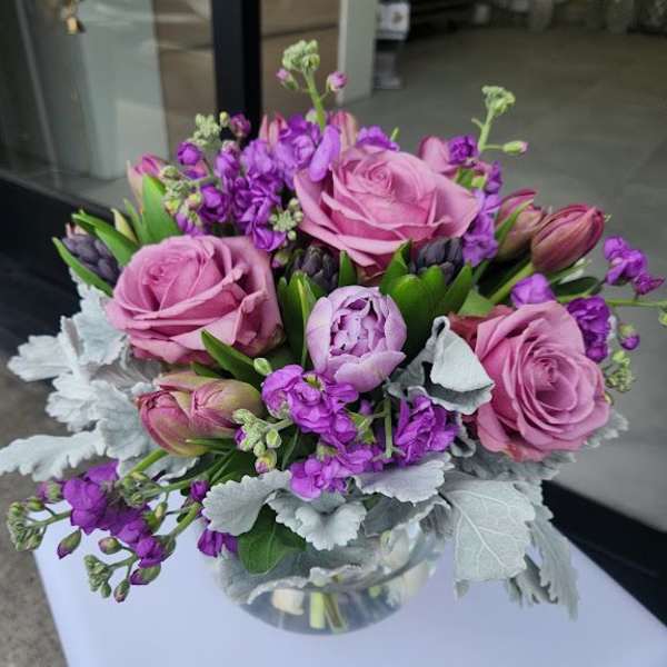 Round arrangement of pink roses and peony with purple blooms in a clear glass bowl vase