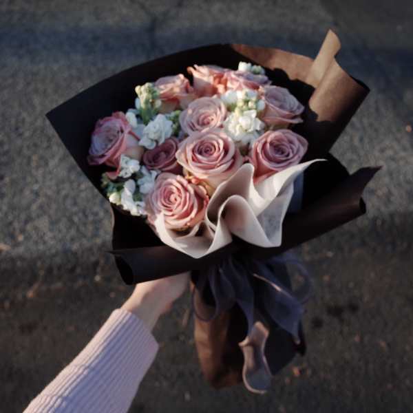 Handheld bouquet of pink roses with white filler flowers wrapped in dark paper