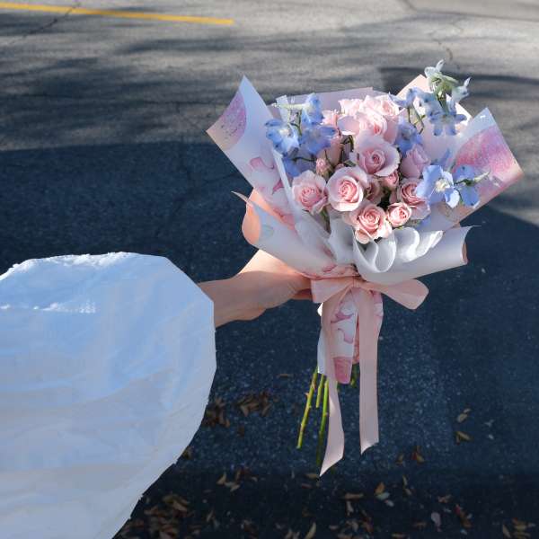 Handheld bouquet of pink roses and pale blue flowers wrapped in pastel paper