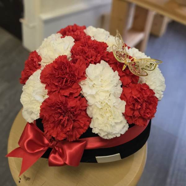 Red and white carnation arrangement in a black hat box with a red ribbon and butterfly decoration