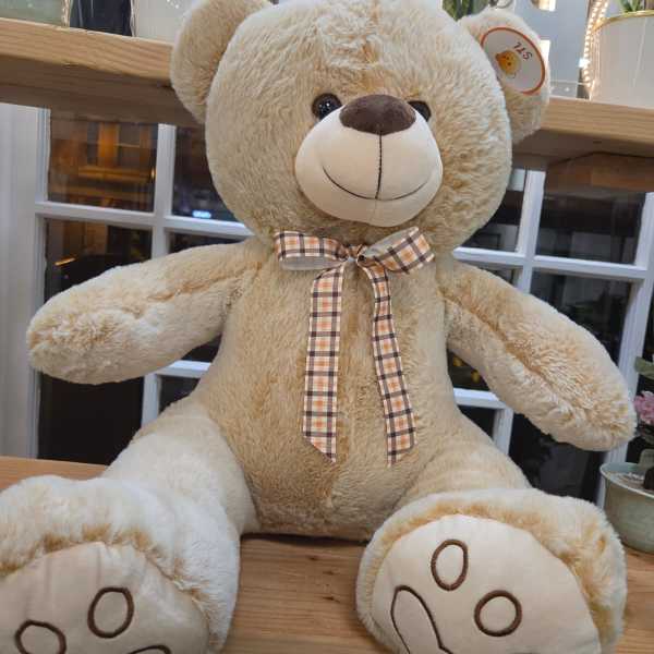 Large beige teddy bear with a plaid bow sitting on a wooden shelf