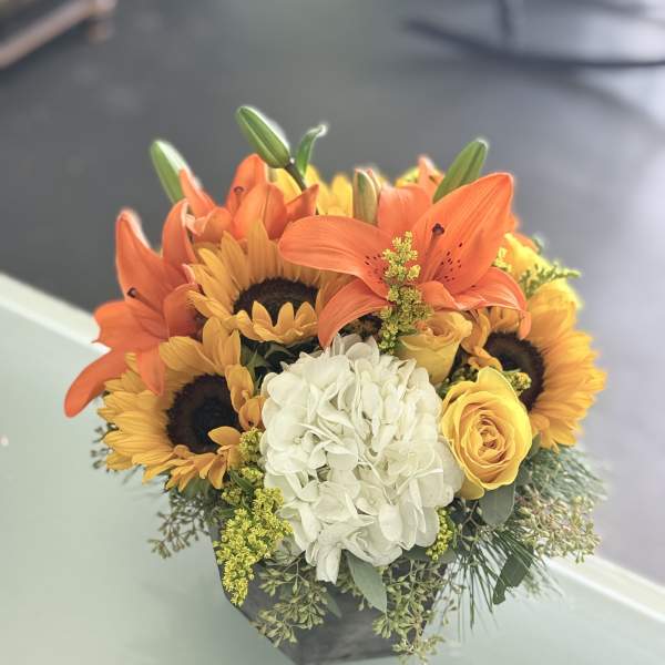 Orange lilies, sunflowers, a white hydrangea, and yellow roses in a low black container.