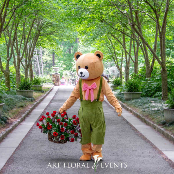 Person in a bear costume carrying a basket of red roses on a tree-lined path