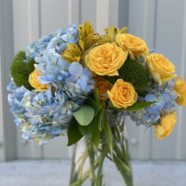Blue hydrangeas and yellow roses arranged in a clear glass vase